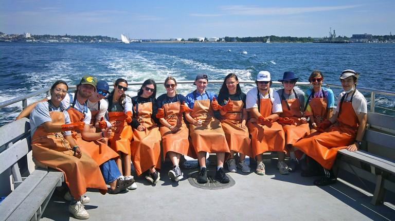 A group of students in fishing apparel sit on a boat in Casco Bay