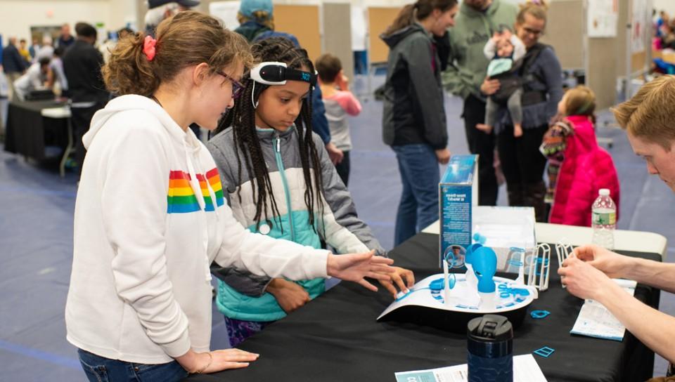 A child wearing scientific equipment learns about its use with her friend at a U N E Brain Fair